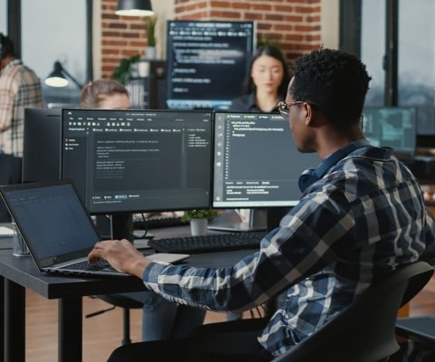 Portrait of system engineer fixing glasses while typing on laptop smiling satisfied about compiling results sitting at desk with multiple screens. App developer enjoying working on user interface.